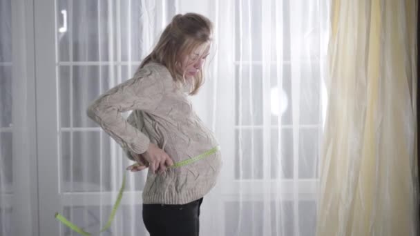 Seitenansicht der schönen kaukasischen schwangeren Mädchen Messen Taillengröße. Aufgeregt werfen sie Maßband weg und unterschreiben. Schwangerschaft, Stimmungsschwankungen, Körperveränderung. — Stockvideo