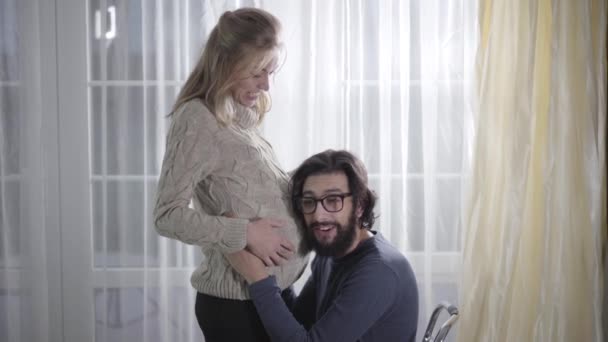 Sidovy över den blonda vita kvinnan och hennes man inomhus. Extremt glad man lyssnar på hans framtida babys hjärtslag. Lyckligt par väntar förlossning. Graviditet och amning. — Stockvideo