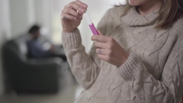 Young Caucasian woman holding pregnancy test as blurred man sitting on sofa at the background. Wife thinking on the way to tell husband about pregnancy. — Stock Video
