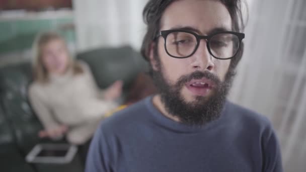 Close-up portrait of young Caucasian man with black curly beard wearing eyeglasses. Blond pregnant woman yelling at him from the background. Pregnancy, gestation, mood swings, patience. — Stock Video