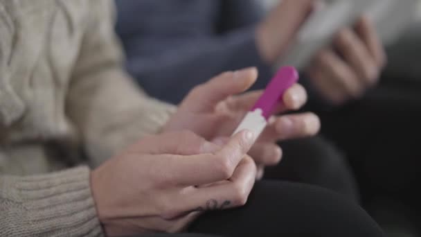 Close-up side view of female Caucasian hands holding affirmative pregnancy test. Blurred male hands at the background using tablet. Wife deciding how to tell husband about pregnancy. — Stock Video
