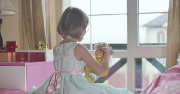 Kaukasisches Mädchen in wunderschönem blauen Kleid mit rosa Schmetterlingen, die am Bett sitzen und mit Puppe spielen. Kind umarmt das Spielzeug und lächelt. Kindheit, Freizeitaktivität. — Stockvideo