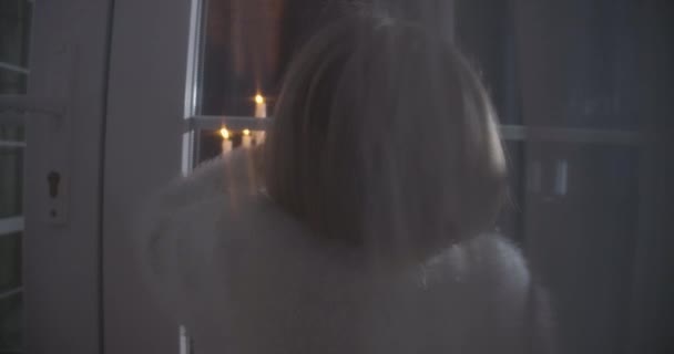 Närbild på baksidan av liten vit blond flicka tittar in i huset genom glasfönster. Ljusstake blixtnedslag inomhus. Skrämsel, rädsla, konstighet. — Stockvideo