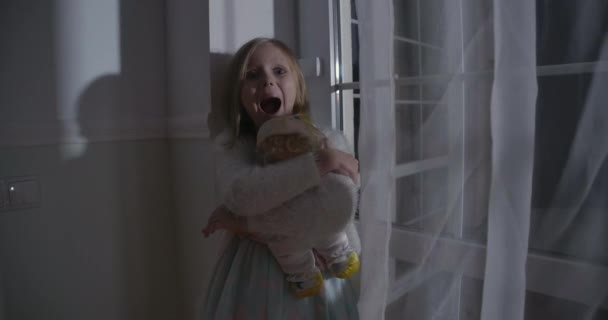 Primer plano de niña rubia caucásica asustada abrazando muñeca y gritando. Retrato de niño asustado de pie junto a la gran ventana con juguete. Miedo, miedo, extrañeza . — Vídeos de Stock