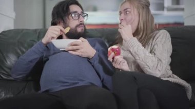 Genç beyaz kadın hamile kocasıyla televizyon izliyor ve tabağından cips yiyor. Sosyal rolleri değişen komik bir aile. Şaka, eğlence, şaka.