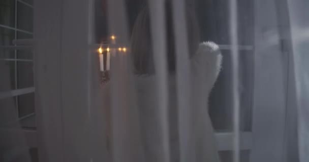 Schießen durch weiße durchscheinende Vorhänge kaukasischer Mädchen, die durch ein Glasfenster ins Haus blicken. Kerzenlicht drinnen. Angst, Angst, Fremdheit. — Stockvideo