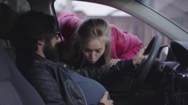 Genç beyaz kadın hamile kocasının karnını öpüyor, eşini öpüyor ve gidiyor. Hamileliği taklit eden adam motoru çalıştırıyor. Sosyal rollerini değiştiren komik bir aile. Şaka, eğlence, şaka.