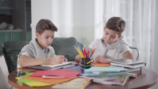 Dva kreativní kavkazští dvojčata sedí u stolu a kreslí barevnými tužkami. Roztomilý chlapec se dívá na fotku svých bratrů a ukazuje palec nahoru. Sourozenci spolu dělají domácí úkoly. Vzdělání. — Stock video
