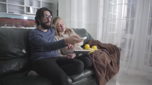 젊은 백인 커플 이 집 소파에 앉아 TV 를 보고 있습니다. 안경을 쓴 남자는 채널을 바꾸고 아름답게 임신 한 아내는 애플을 먹고 태블릿 화면을 보고 있다. 임신 중절. — 비디오