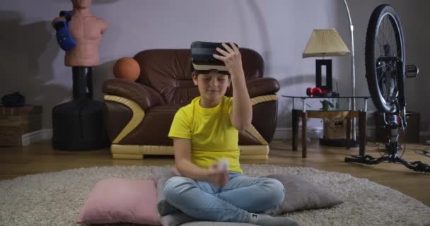 Retrato de un adolescente caucásico que toma los auriculares VR y usa control remoto para activar el juego. Niño probándose un dispositivo en casa. Auriculares virtuales, juegos en línea, realidad 3d. Sede del cine 4k ProRes . — Vídeo de stock