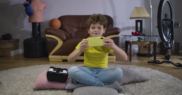 행복 한 백인 십 대 남자 아이가 집의 바닥에 앉아 비디오 게임을 하고 있습니다. 스마트폰을 사용하여 곱슬머리를 하고 앞을 바라보고 있는 쾌활 한 소년. 온라인 게임, 기술. 영화 4K 전립선 HQ. — 비디오