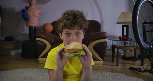Elnyelt kaukázusi tinédzser fiú TV-t néz, hamburgert rágcsál, köhög. Aranyos tinédzser gyerek portréja gyorskajával otthon. Szabadidős tevékenység, kikapcsolódás, generáció Z. Cinema 4k Prores Hq. — Stock videók