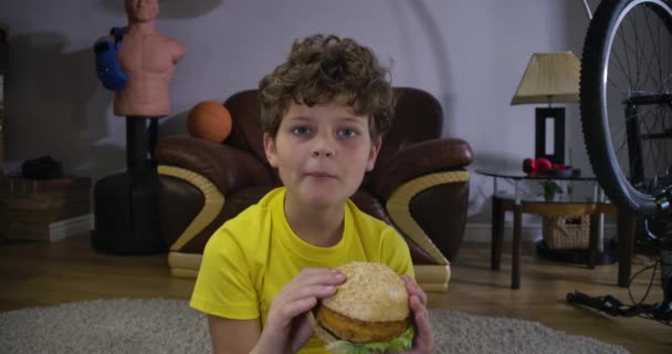 Gesicht eines kaukasischen, lockigen Jungen, der Hamburger kaut und fernsieht. Netter Teenager mit Fast Food zu Hause. Freizeitaktivität, Entspannung, Generation z. Kino 4k prores hq. — Stockvideo