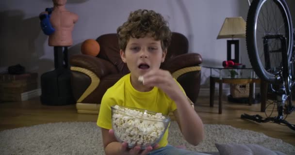 Portrait d'un petit garçon caucasien regardant la télévision et mâchant du pop-corn. Mignon adolescent aux cheveux bouclés mangeant de la malbouffe à la maison. Loisirs, détente, génération Z. Cinema 4k ProRes HQ . — Video