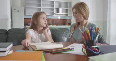 Genç, güzel, beyaz bir anne kızına ödev yaptırıyor. Üzgün çocuk çalışmayla ilgili zorluklar yaşıyor. Ebeveyn çocuğuna derslerinde yardım ediyor. Eğitim, zeka. Sinema 4k Prores Merkezi.