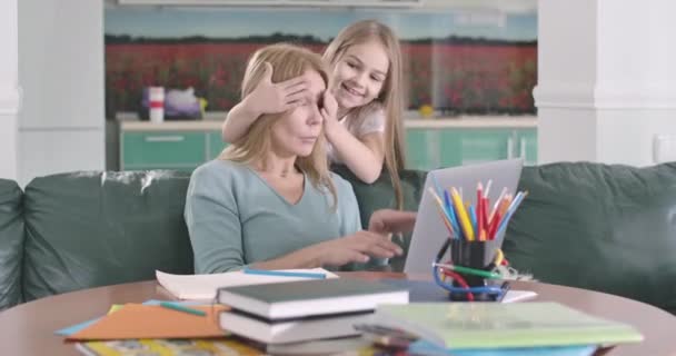 Portret van een serieuze blanke vrouw die werkt met een laptop als haar blije dochter die van de achtergrond komt en haar ogen sluit met handen. Een vrouw die het kind knuffelt. Bioscoop 4k Prores Hq. — Stockvideo