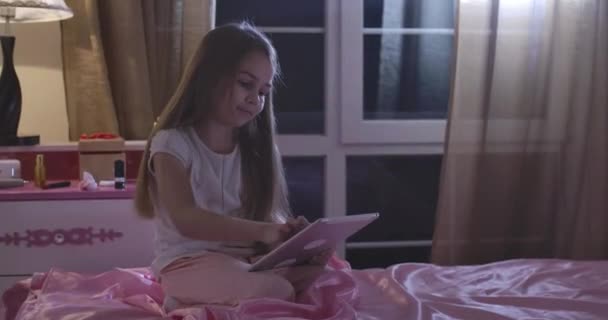 Porträt einer charmanten kaukasischen Schülerin, die auf einem rosafarbenen Bett sitzt und ein Tablet benutzt. Teenagermädchen nutzt abends soziale Medien. Internetsucht, Freizeit, Nachrichten. kino 4k prores hq. — Stockvideo