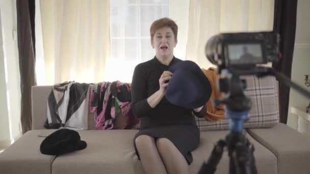 正面的白人妇女的画像，带着相机帽和围巾。 一个成熟的女人坐在沙发上，为互联网页面录制视频。 社交媒体、技术. — 图库视频影像