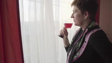 Düşünceli, kendine güvenen beyaz iş kadını kırmızı şarap içiyor ve akşamları pencereden dışarı bakıyor. Bir kadeh şarapla ayakta duran güzel bir kadın. Kendine güven, dinlenme. Boş zaman.