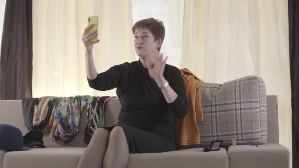 Πορτρέτο της όμορφης καυκάσιας ηλικιωμένης κυρίας που καταγράφει βίντεο selfie στο smartphone της. Ώριμη γυναίκα μπλόγκερ χαιρετά συνδρομητές, βάζει το τηλέφωνο στον καναπέ και αναστενάζει. Σύγχρονη ζωή, blogging. — Αρχείο Βίντεο