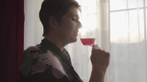 Detailní záběr usmívající se bělošky popíjející červené víno a dívající se z okna. Portrét sebevědomé dospělé dámy odpočívající ve večerních hodinách. Sebevědomí, odpočinek. volný čas. — Stock video