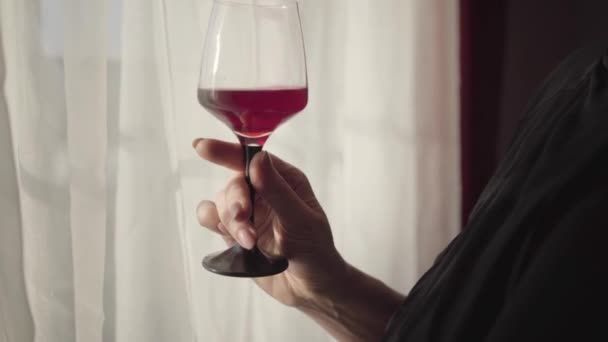 Nahaufnahme einer eleganten weiblichen kaukasischen Hand, die ein Glas mit Rotwein hält. Frau mit stilvoller Maniküre mit Weinglas im Hintergrund des Sonnenlichts, das durch den weißen Vorhang scheint. — Stockvideo
