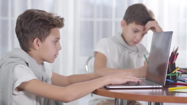 Vista lateral de primer plano de chico caucásico jugando juegos en línea en el ordenador portátil. Su hermano gemelo sentado en el fondo, haciendo los deberes. Diferente actitud hacia el estudio, concepto de educación, adicción a Internet . — Vídeos de Stock