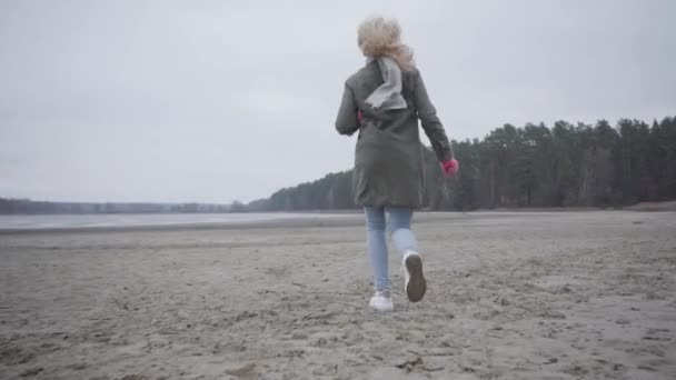 Kamera sledující dospělou blondýnu bělošku v teplých šatech běžící na podzimní pláži. Radostná dívka se baví navzdory počasí venku. — Stock video