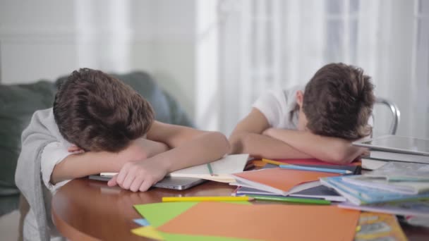 Ung vit brunett pojke vakna upp och stretcha. Hans bror eller vän sover på bordet. Syskon somnade på böcker efter läxorna. Utmattning, utbildningskoncept. — Stockvideo