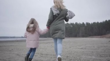 Sonbahar plajı boyunca koşan beyaz kadın ve küçük kızın arka görüntüsü. Neşeli Kafkas anne ve kızı dışarıda eğleniyor..