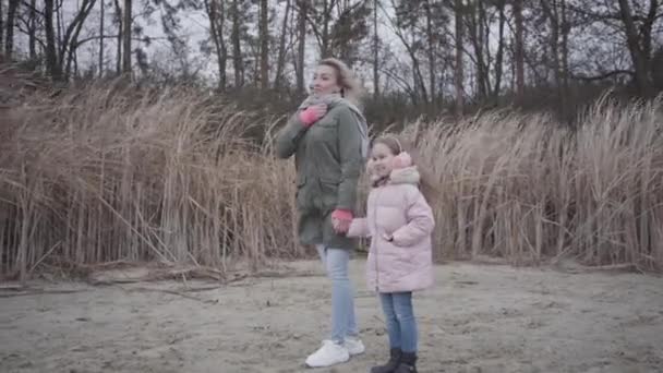 Side udsigt over smilende kaukasiske kvinde og munter pige stående ved efteråret stranden og ser væk. Familie venter på nogen udendørs . – Stock-video