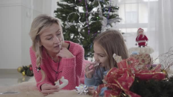 Portret van een blanke moeder en dochter die op de grond liggen en koekjes eten. Vreugdevolle moeder die haar kind hoort praten. Gelukkig gezin rustend op de achtergrond van de kerstboom. — Stockvideo