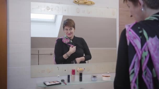 Mature Caucasian senior woman looking at two scarfs in front of big mirror in bathroom. Elegant businesswoman choosing outfit in the morning. Style, fashion, elegance. — Stock Video