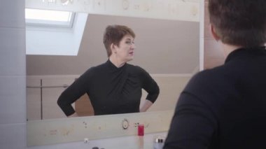 Güzel esmer, olgun bir kadın büyük aynaya bakıyor ve yüz kremlerini kokluyor. Makyaj yapan beyaz bir kadın. Moda, güzellik, yüz bakımı, cilt bakımı.