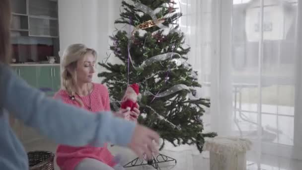 Baksidan av lilla vita flicka hjälper mamma att dekorera julgran. Vacker blond vit kvinna och hennes brunett dotter förbereder sig för jul. Familjeinredning hem för fest. — Stockvideo