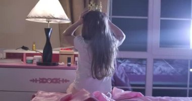 Kafasında oyuncak taçla pembe yatakta oturan Kafkasyalı genç kızın arkası. Okul çocuğu yatmaya hazırlanıyor da. Çocukluk, dinlenme, uyku zamanı. Sinema 4k Prores Merkezi.