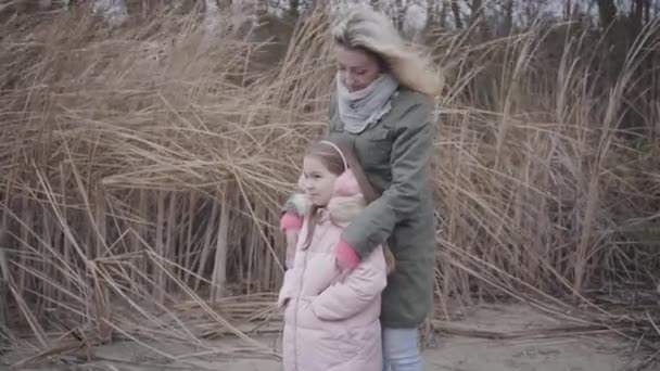 Pohled z boku krásné bělošky a její dcery oblečené v růžových šatech, stojící ve větru a dívající se stranou. Matka drží holčičku a mluví. Volný čas, volný čas, podzimní aktivity. — Stock video