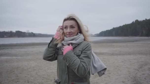 Cámara acercándose a la hermosa mujer rubia caucásica de pie frente al lago o río de otoño. Muchacha atractiva con ojos marrones sosteniendo el pelo en el viento y hablando . — Vídeos de Stock