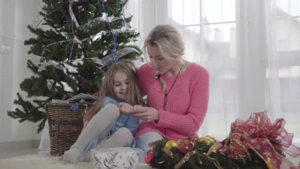 Sonriente chica caucásica sentada con una mujer adulta en el fondo del árbol de Navidad. Madre hablando con su hijo adentro. Familia preparándose para la celebración de Año Nuevo . — Vídeos de Stock