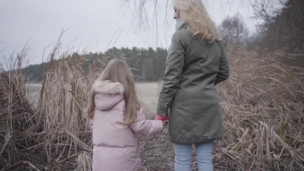 Rückansicht einer erwachsenen blonden Frau und eines kleinen brünetten Mädchens, die in herbstlichen Büschen vor einem See oder Fluss stehen. Mutter und Tochter ruhen sich im Freien aus. Familie, Freizeit, Zweisamkeit. — Stockvideo