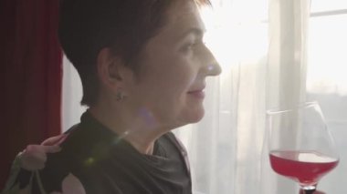 Güneş ışınlarında şarap kadehiyle dikilen mutlu, beyaz bir kadının yan görüntüsü. Esmer, olgun bir bayan akşamları pencereden dışarı bakar ve gülümser..