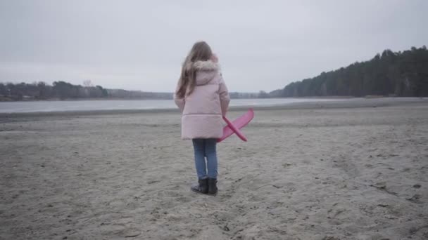 Achteraanzicht van een blank schoolmeisje in roze kleren die in de wind staat op de rivieroever en een speelgoedvliegtuig vasthoudt. Kind dat alleen buiten wacht. — Stockvideo