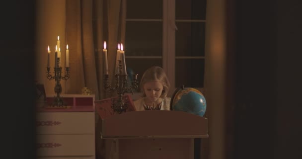 Mała biała uczennica siedzi przy biurku i odrabia lekcje przy świecach. Blond dziecko słyszy coś przerażającego i rozgląda się przerażająco. Samotność, strach, strach. — Wideo stockowe