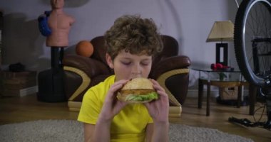 Televizyonun önünde hamburgeri ısıran beyaz çocuğun yakın plan yüzü. Genç kıvırcık saçlı çocuk evde film izliyor. Boş zaman aktivitesi, rahatlama, Z. Cinema 4k Prores Hq jenerasyonu..