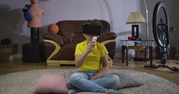 VR 구글의 코카서스 소년 이 리모컨을 들고 구부리고 있습니다. 노란 티셔츠와 파란 청바지를 입은 학생은 집에서 증강현실을 사용 합니다. 3D 현실, 가상 현실. 영화 4K 전립선 HQ. — 비디오