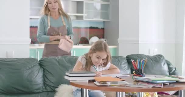 Portret van een blank schoolmeisje dat huiswerk maakt aan tafel terwijl haar moeder van de achtergrond komt. Schattig kind en volwassen vrouw lachend en knuffelend. Onderwijs, studeren. Bioscoop 4k Prores Hq. — Stockvideo