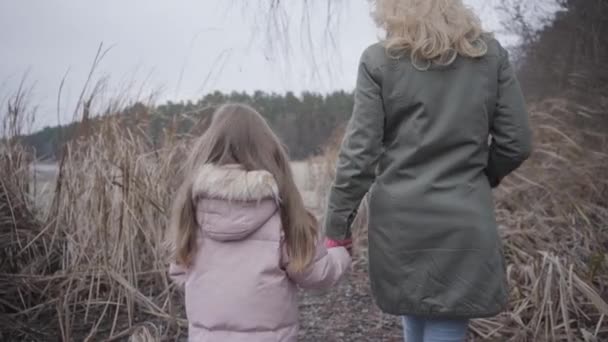 Pohled zezadu na bělošku a dívku kráčející k jezeru nebo řece křovím. Blond matka a brunetka dcera odpočívají venku. Rodina, volný čas, spolužití. — Stock video