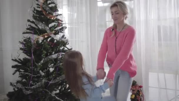Портрет молодої кавказької матері, що тримає дочок за руки перед різдвяним деревом. Жінка готується до відпустки зі своєю дитиною. Родина прикрашає дім для святкування.. — стокове відео
