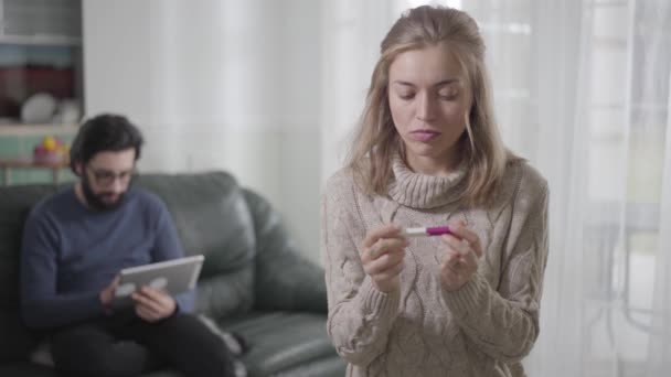 Portrait of young Caucasian blond woman holding affirmative pregnancy test, looking back at her husband, and thinking. Beautiful sad girl upset about pregnancy. — Stock Video