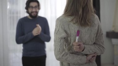 Arkasında iki rütbe olan beyaz bir kadının hamilelik testi tutuşu. Gözlüklü, gülümseyen koca arka plandaki şaşkınlığı soruyor..